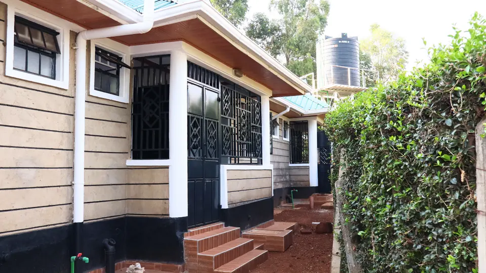 Side of a bungalow with steps to its veranda, a hedge and a raised water tank behind